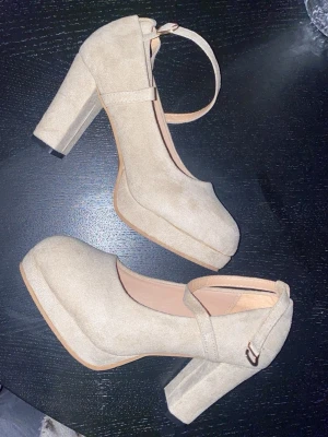 Beige platåpumps med blockklack - Snygga beige pumps med hög blockklack och platåsula. Skorna har en rundad tå och justerbart ankelband med spänne. Tillverkade i mockaimitation för en trendig look. Perfekta för dig som vill ha både höjd och stabilitet. Oanvända!