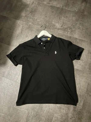 Svart pikétröja från Polo Ralph Lauren - Klassisk svart pikétröja från Polo Ralph Lauren med vit broderad logga på bröstet. Tröjan har korta ärmar, krage och knappar framtill. Perfekt till jeans och nu emot sommaren.