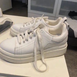 Vita sneakers med platåsula - Säljer ett par helvita sneakers med platåsula och snörning. Skorna har rund tå och är tillverkade i syntetmaterial med tydliga sömmar och klassisk design. Perfekta för dig som gillar en clean och stilren look.