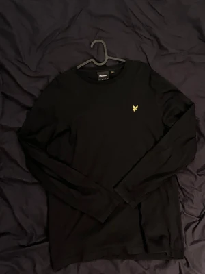 Svart långärmad tröja Lyle & Scott - Snygg svart långärmad tröja från Lyle & Scott med rund halsringning och den klassiska gula logon broderad på bröstet. Tillverkad i mjukt bomullsmaterial som känns skönt mot huden. Perfekt basplagg för en clean och stilren look.