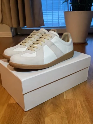 Maison marigela gata - Snygga sneakers i vitt och beige från Maison Margiela med klassisk siluett. Skorna har ovandel i läder och mocka, beigea snören och en brun gummisula. Perfekta för dig som gillar stilrena och trendiga sneakers med retrovibbar.