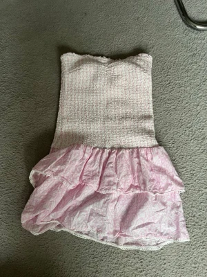 Rosa axelbandslös volangklänning - TRYCK GÄRNA KÖP DIREKT💗Söt axelbandslös kortklänning i ljusrosa med smockad överdel och dubbla volanger nertill. Klänningen har ett diskret mönster och är perfekt för varma dagar. Materialet känns lätt och luftigt💗