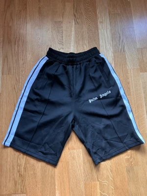 Palm Angels svarta shorts med vita ränder - Svarta shorts från Palm Angels med vita sidoränder och logga tryckt på vänster ben. De har elastisk midja och sportig look, perfekta för en avslappnad streetwear-stil.