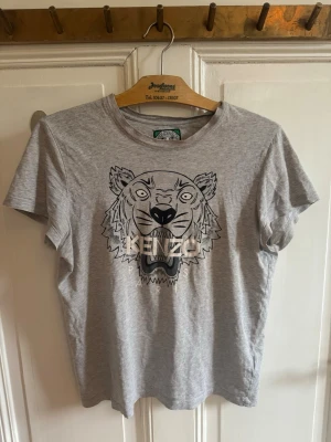 Grå Kenzo t-shirt med tigertryck - Snygg grå t-shirt från Kenzo med ikoniskt tigertryck och logga på bröstet. Klassisk passform och rund halsringning. Tillverkad i mjuk bomull som känns skön mot huden. Perfekt för dig som vill sticka ut med en stilren men cool look. Defekter syns på baksidan men snygg på!!