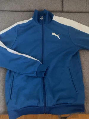 Sportswear, Träningsjacka - Puma - Snygg blå träningsjacka från Puma med vita ränder längs ärmarna och dragkedja framtill. Jackan har ståkrage, muddar vid ärmslut och nederkant samt två fickor. Puma-logga i vitt på bröstet. Perfekt för en sportjacka. OBS: Dragkedjan kan vara lite klurig men det funkar efter nån sekund. (Gammal men är i bra skick för sin ”ålder”).