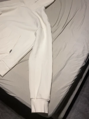 Vit hoodie med huva - Snygg vit hoodie med huva och magficka. Klassisk modell med ribbade muddar vid ärmslut och nederkant. Perfekt för en clean och stilren look. Mjuk och skön bomullskänsla, passar till det mesta i garderoben.