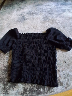 Svart smockad topp från WE - Trendig svart topp från WE med smockad kropp och puffiga korta ärmar. Toppen är elastisk och har en croppad passform som ger en cool och modern look. Perfekt att matcha med höga jeans eller kjol.