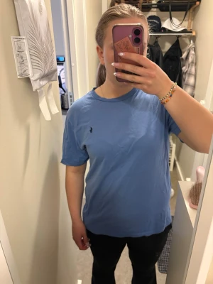 Blå t-shirt från Polo Ralph Lauren - Blå t-shirt från Polo Ralph Lauren med klassisk rund hals och broderad logga på bröstet. T-shirten är kortärmad och har en avslappnad passform. Perfekt för en stilren och enkel look. Storleken är barn xl/170 och är från kidsbrandstore