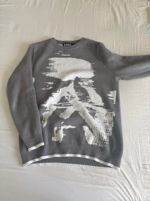 Grå tröja med vit grafisk print SOUL - Grå långärmad tröja från SOUL med en stor vit grafisk print framtill som ger en modern och edgy look. Rund halsringning och ribbade muddar vid ärmslut och nederkant. Perfekt för dig som gillar streetwear och unika prints.