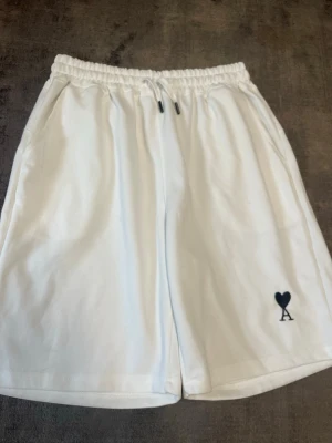 Vita shorts från Ami Paris med hjärtdetalj - Snygga vita shorts från Ami Paris med elastisk midja och snörning. På vänster ben finns en svart hjärtdetalj med bokstaven A. Perfekta för sommaren och har en avslappnad passform.