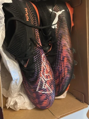 Puma Future Z fotbollsskor svart/lil - Snygga Puma Future Z fotbollsskor med svart bas och coolt mönster i lila, orange och vitt. Skorna har stickad strumpliknande krage för extra stöd och greppvänlig ovandel i syntetmaterial. Perfekta för dig som vill sticka ut på planen! Helt 
