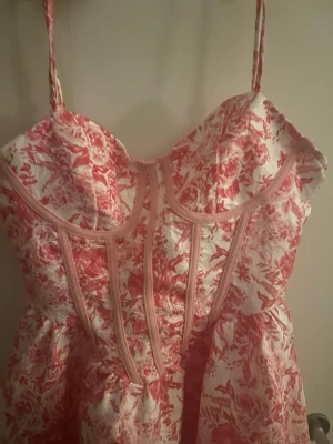 Rosa och vit blommig klänning - Söt klänning med smala axelband och blommigt mönster i rosa och vitt. Klänningen har markerade sömmar framtill som ger en korsettliknande look och en lätt utställd kjol. Perfekt för dig som gillar romantisk och feminin stil. Funkar för utekväll, fester och födelsedagar och passar för student veckan. Storlek 34/36