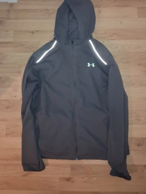 Grå vindjacka från Under Armour - Säljer min under armour windbreaker använt den ett fåtal gånger, bra för sommaren. OBS finns ett litet hål i baksidan skriv om ni vill se den annars är den perfekt.