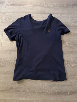 Mörkblå t-shirt från Ralph Lauren - Mörkblå t-shirt från Ralph Lauren med klassisk passform och korta ärmar. T-shirten har en liten broderad logga på bröstet och är tillverkad i mjuk bomull. Perfekt för en stilren och enkel look.