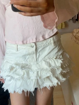 Vit volangkjol från Monki - Supersöt vit kortkjol från Monki med flera lager volanger och råa kanter för en trendig look. Kjolen har bälteshällor och knapp framtill. Perfekt för dig som vill sticka ut med en lekfull och edgy stil. Strl 34