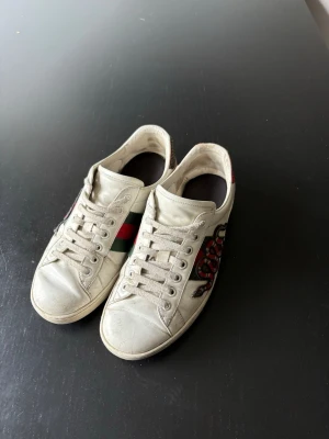 Gucci ace 🐍 - Säljer ett par vita Gucci sneakers med klassiska gröna och röda ränder på sidan samt en cool broderad orm på andra sidan. Skorna är låga och har snörning framtill. Materialet är läder och designen är riktigt trendig.