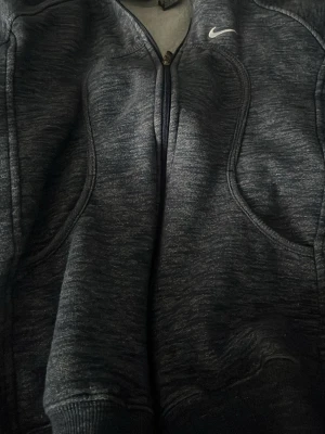 Mörkgrå zip hoodie från Nike - Snygg mörkgrå zip hoodie från Nike med dragkedja framtill och två fickor. Hoodien har en liten vit Nike-logga på bröstet och en klassisk huva. Tillverkad i mjukt och bekvämt material, perfekt för chill dagar eller träning.