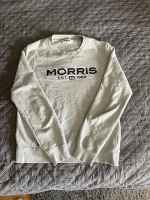 Grå sweatshirt från Morris - Snygg grå sweatshirt från Morris med svart broderad logga och texten 'MORRIS EST. 1968' framtill. Tröjan har rund halsringning, långa ärmar och ribbade muddar vid ärmslut och nederkant. Perfekt för en avslappnad och stilren look.