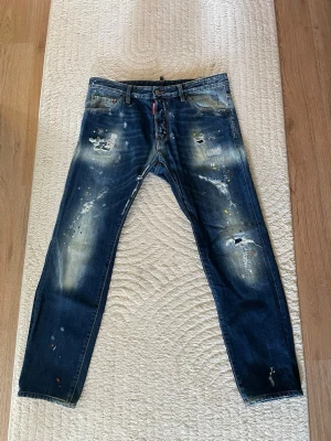 ÄKTA Dsquared2 jeans - Säljer ett par mörkblå jeans från Dsquared2 med slitna detaljer, patchar och färgstänk. Jeansen har rak passform, klassiska fem fickor och tydliga slitningar framtill för en cool, trendig look. Perfekta för dig som gillar streetstyle och unika plagg.
