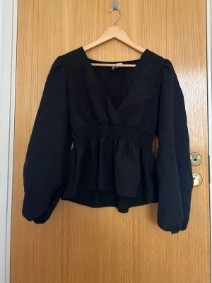 Svart omlottblus från H&M Divided - Snygg svart omlottblus från H&M Divided med markerad midja och långa puffärmar. Blusen har v-ringning och smockdetalj i midjan som ger en trendig siluett. Perfekt att styla med jeans eller kjol för en cool look.