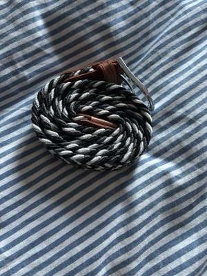 Svartvitt flätat bälte med läderdetalj - Snyggt flätat bälte i svart och vitt med bruna läderdetaljer och silverfärgat spänne. Perfekt accessoar för att lyfta din outfit och funkar till både jeans och byxor. Bältet har en flexibel design som gör det enkelt att justera längden.