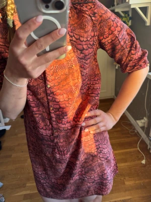 Röd mönstrad kortklänning - Kortklänning i röd och brun ton med unikt mönster som påminner om fjäll eller skala. Klänningen har knappar framtill, trekvartsärm och avslappnad passform. Perfekt för dig som gillar statementplagg och vill sticka ut.