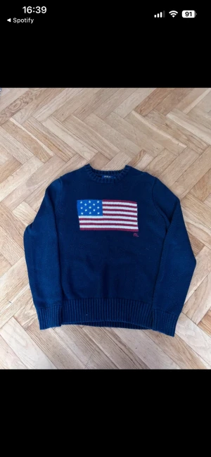 Mörkblå stickad tröja Polo Ralph Lauren - Mörkblå stickad tröja från Polo Ralph Lauren med amerikansk flagga broderad på bröstet och RL-detalj. Tröjan har rund halsringning, ribbade muddar och nederkant. Perfekt för dig som gillar klassisk preppy stil.