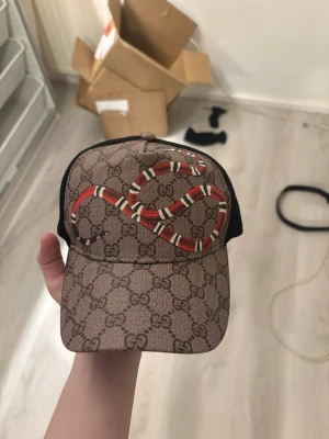 Beige keps - Svart premiumkeps med diskret monogrammönster och iögonfallande röd orm-design på framsidan. Modern trucker-modell med mesh baktill för skön passform och ventilation. Passar perfekt till både vardag och streetwear-stil.