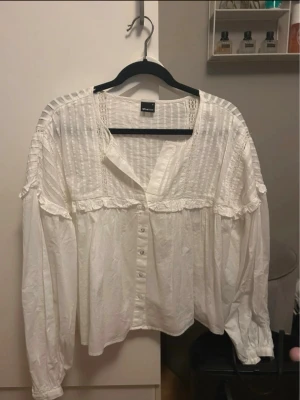 Vit blus från Gina - Söt vit blus från Gina med knappar framtill, volangdetaljer och diskret rutmönster upptill. Blusen har lång ärm med resår vid ärmslut och en lös, luftig passform. Perfekt för en trendig och avslappnad look.