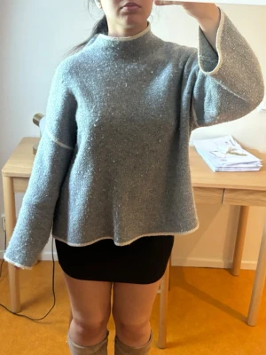 Zara tröja  - En grå stickad tröja med vita detaljer längs kanterna och sömmarna. Tröjan har en hög hals och långa ärmar, perfekt för att hålla sig varm under kyliga dagar. Passformen är loose och materialet känns mjukt och bekvämt.