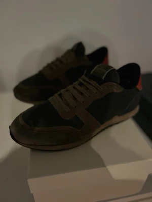Valentino sneakers i grönt och brunt - Snygga sneakers från Valentino i grönt tyg med bruna mockadetaljer och beige sula. Skorna har svarta och röda detaljer vid hälen samt klassisk snörning. Perfekta för dig som vill ha en stilren men ändå unik look.vid snabb affär 600kr 