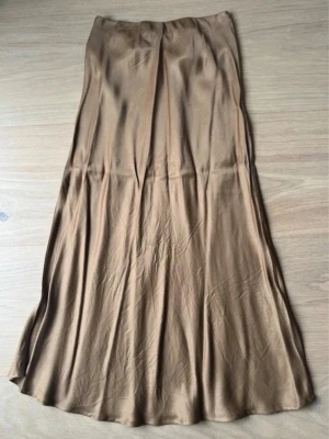 Beige satinkjol i lång modell - Säljer en snygg beige långkjol i satinliknande material. Kjolen har en lätt glansig finish och faller mjukt längs kroppen. Perfekt för dig som gillar en stilren och enkel look. Passar till många olika outfits och är superenkel att styla.