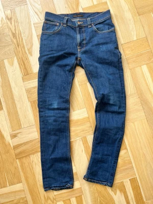 Mörkblå skinny jeans från Nudie - Säljer ett par mörkblå skinny jeans från Nudie med klassiska kontrastsömmar och broderade bakfickor. Jeansen har fem fickor, normal midja och är gjorda i stretchig denim för skön passform. Perfekta för dig som gillar en smal siluett.
