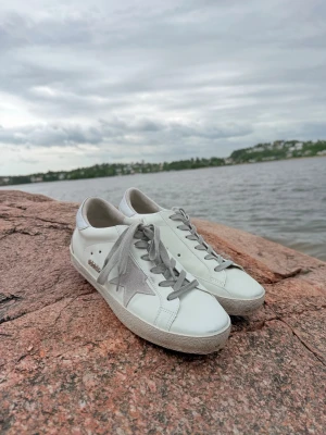 Golden Goose vita sneakers med grå stjärna - Snygga vita sneakers från Golden Goose med grå stjärna på sidan och grå skosnören. Skorna har en klassisk låg modell och är tillverkade i läder med slitstark gummisula. Perfekta för en trendig och avslappnad stil.