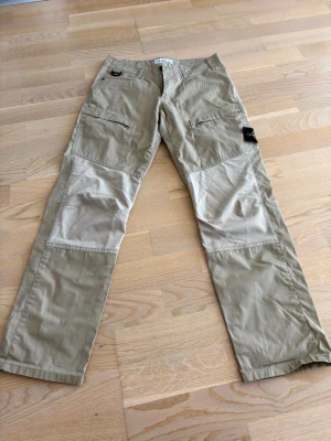 Beige cargobyxor stone Island  - Snygga cargobyxor i beige med kontrasterande ljusare partier på knän och bak. Byxorna har flera fickor med dragkedja och en klassisk cargomodell. Perfekta för en avslappnad streetstil.