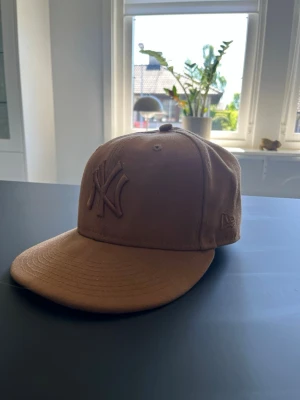 Beige New Era NY keps - Skärmen är knäckt men de ska inte påverka funktionen. Snygg beige keps från New Era med broderad NY-logga framtill. Klassisk modell med böjd skärm och ventilerande hål upptill. Perfekt accessoar för en avslappnad streetwear-look.
