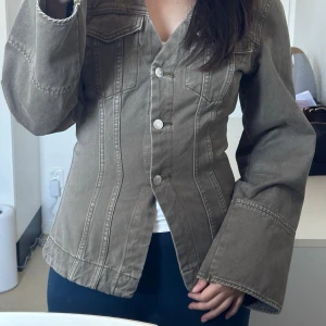 Olivgrön shaped denim jacket från GinaTricot - olivgrön jeansjacka med markerade sömmar och utsvängda ärmar. Jackan har V-ringning, knäppning framtill med silverfärgade knappar 🫒helt ny, aldrig använd🔥