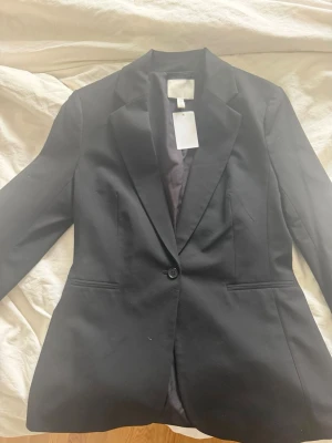 Svart kavaj med klassisk passform - Stilren svart kavaj med enknäppning och figursydd siluett. Klassisk krage och två fickor framtill. Perfekt för en snygg och uppklädd look. Materialet är slätt och fodrat för extra komfort.