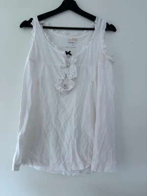 Vit blus med volang från Odd Molly - Supersöt vit blus från Odd Molly med volangdetaljer och knappar framtill samt en liten svart rosett vid halsen. Blusen är ärmlös och har en romantisk känsla med fina ryschkanter. Perfekt att styla till jeans eller kjol.