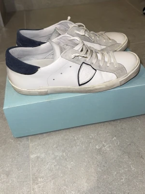 Philippe Model vita sneakers med blå detalj - Snygga vita sneakers från Philippe Model med grå mockadetaljer och mörkblå häl. Klassisk låg modell med vita snören och logotyp på sidan. Skorna har en stilren design och är tillverkade i läder och mocka. Perfekta för en trendig och avslappnad look.