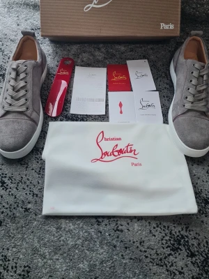 Gråa Christian Louboutin sneakers - Snygga gråa sneakers från Christian Louboutin i mocka med klassisk röd sula och vita kanter. Lågt skaft, rund tå och matchande grå skosnören. Kommer med originalkartong, dustbag och tillbehör. Perfekta för dig som vill sticka ut med exklusiva detaljer.