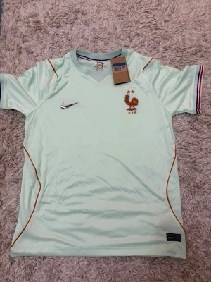 Frankrike fotbollströja  ljusgrön - Snygg ljusgrön fotbollströja från Nike med Frankrikes landslagsemblem och två stjärnor på bröstet. Tröjan har orange detaljer och broderad Nike-logga. Ärmarna har randiga muddar i blått, vitt och rött. Perfekt för fotbollsträning eller match.