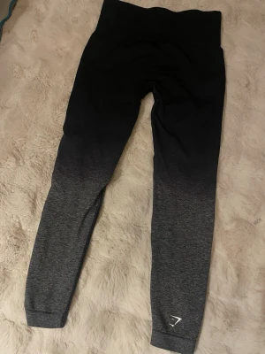 Svarta och grå ombre leggings Gymshark - Snygga leggings från Gymshark i svart och grått ombre-mönster. Hög midja och stretchigt material som sitter skönt och formar sig efter kroppen. Perfekta för träning eller chill. Liten Gymshark-logga vid benet.