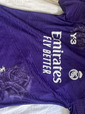 Lila Real Madrid x Y-3 t-shirt - Cool lila t-shirt från Y-3 och Real Madrid med vita loggor och text. Unikt blommönster i form av rosor på framsidan. Perfekt för dig som vill sticka ut med sportig stil och exklusivt samarbete.