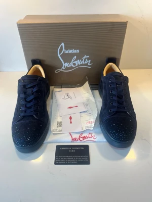 Christian Louboutin blåa sneakers - Snygga mörkblå sneakers från Christian Louboutin med klassiska röda sulor och diskreta nitar på tån. Skorna har en låg profil, snörning och är tillverkade i lyxigt mockamaterial. Perfekt för dig som vill sticka ut med exklusiva detaljer och ikonisk design.