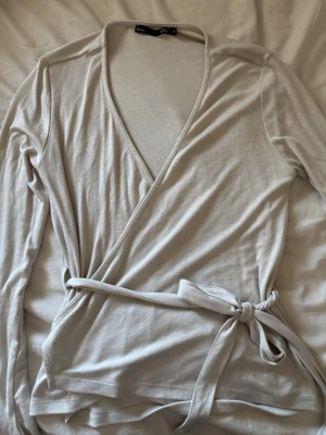 Beige omlottopp med knytband - Snygg beige omlottopp från Gina Tricot med långa ärmar och knytband i midjan. Toppen har en djup v-ringning och är tillverkad i mjuk bomullsmix som sitter skönt mot kroppen. Perfekt för en stilren och enkel look. 