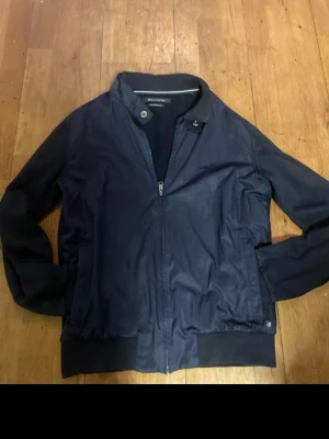 Marinblå cardigan Marc O'Polo - Snygg marinblå bomberjacka/ cardigan från Marc O'Polo med dragkedja framtill och ribbade muddar vid ärmslut och midja. Jackan har en klassisk bomberkrage med knappdetalj och är tillverkad i ett mjukt material som kallas carbonised sweat.