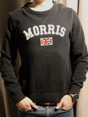 Svart tröja från Morris med tryck - Snygg svart tröja från Morris med vit text och brittiska flaggan broderad på bröstet. Tröjan har rund halsringning och ribbade muddar vid ärmslut och nederkant. Perfekt för dig som gillar klassisk och stilren design.