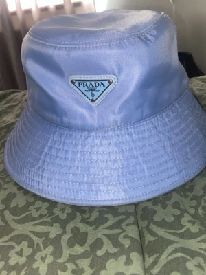 Ljusblå Prada hat , möts bara upp i Stockholm  - Snygg ljuslblå bucket hat från Prada med klassisk triangelformad logotyp framtill. Hatten är tillverkad i glansigt nylonmaterial och har bred brätte med sömdetaljer. Perfekt för att lyfta din streetstyle-look.
