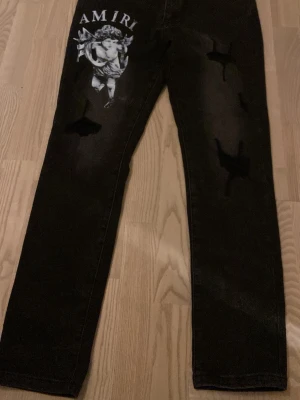 Svarta Amiri jeans med ängeltryck - Säljer ett par svarta jeans från Amiri med ett stort vitt ängeltryck och texten 'AMIRI' på ena benet. Jeansen har raka ben och slitna detaljer på framsidan för en edgy look. Materialet är klassisk denim som känns både robust och trendigt.